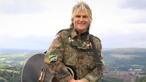 Mike stood in the countryside, you can see the hills behind him. He is holding a guitar with the strap around his shoulder and an army jacket on top. The man is The Alarm's Mike Peters