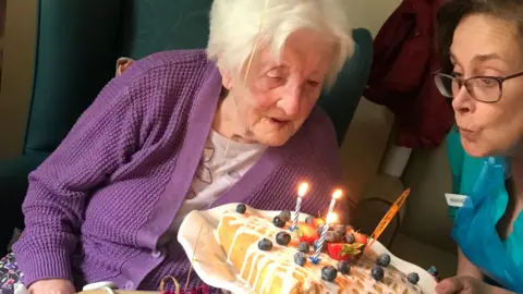 The Mill House Care Home Vera, wearing a purple cardigan blows out several candles on top of her birthday cake along with a carer wearing a green uniform and a blue apron.