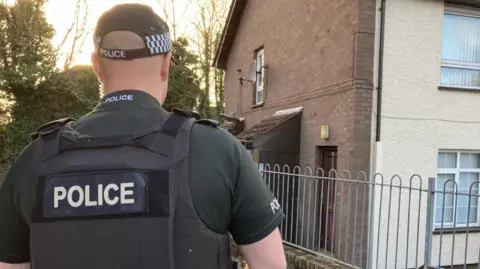 BBC Shows a police officer with their back to the camera near a bricked house with silver railing.