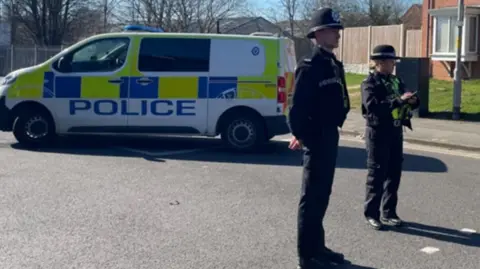 BBC A police van on a residential road with a police man and police woman standing in front of the vehicle.