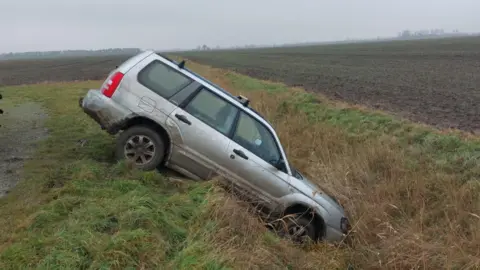 Cambridgeshire Police A silver car has nosedived into a ditch. The back wheels are at the top of a mound of grass and the front of the car is touching the ground. The surrounding area looks to be fields used for farming. 