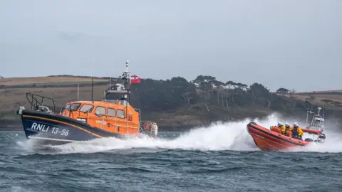 Two RNLI lifeboats moving on the water. On the smaller lifeboat, to the right, there are coastguards on board. Both lifeboats are orange. 