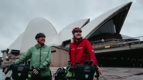 Family Handout Josh and George Kohler and standing behind bicycles in front of the Sydney Opera House, which is a large building that has an obscured shape white roof. They are both reading helmets and coats.