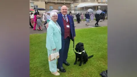 John Hardy John Hardy with his arm around his wife's shoulders. They are both in suits standing in the garden of Buckingham Palace. John is holding Sidney, a black Labrador, on a lead.