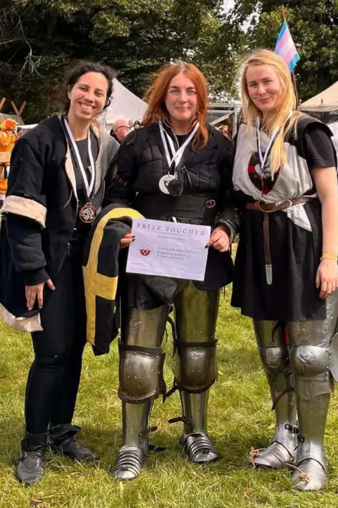 Mary George At an outdoor festival setting, three women stand shoulder to shoulder on grass. Each of them wears a black jacket over their armour. Medals hang from ribbons around their necks. Jasmine holds a rectangular paper certificate that reads “Prize Voucher” at the top. In the background are leafy trees, pale canvas tents, wooden display frames. 