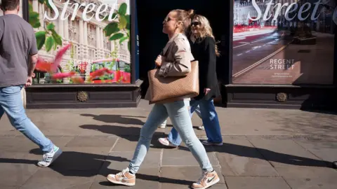 A woman in jeans and carrying a large tote bag walking along a high street. She is seen in profile against the backdrop of a shop window in London and other shoppers are in the background