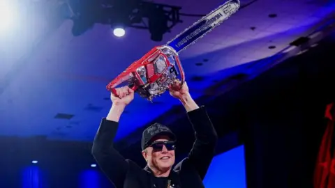 Reuters Elon Musk wearing a blue cap and large dark glasses with a dark-coloured top. He is holding a red chainsaw above his head. He is on a stage with blue ceiling and flats.