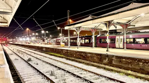 walking tractor/WEATHERWATCHERS Norwich Station. It is dark. In the foreground there are two icy railway lines. The platforms are empty. On the right, next to a platform, there is a long, empty train. 