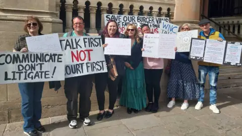 A group of around eight people stand outside a stone building with columns, holding handmade protest signs. The signs include messages such as “Communicate don’t litigate!”, “Talk first not court first,” and “Save our burger vans".