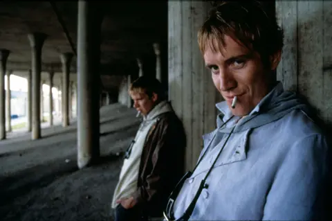 Moviestore Collection Ltd Two men with blond hair both smoking a cigarette standing underneath a motorway flyover with pillars in the background holding up the road deck