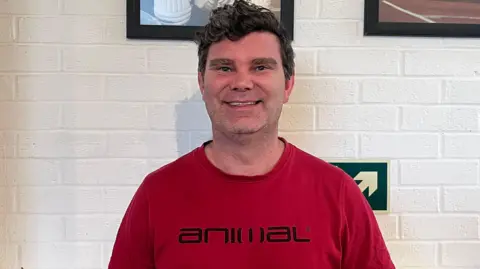 A dark-haired man in a red T-shirt which has a black Animal logo on the front. He is smiling and standing in front of a white brick wall inside a club house, with black-framed photos of athletes on the wall behind him.
