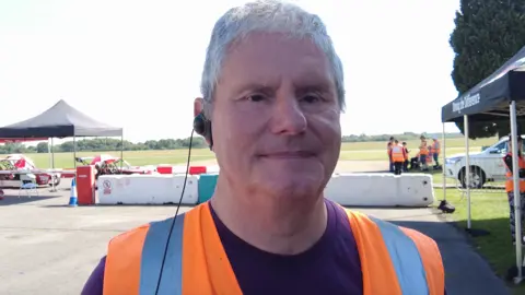 Mike Newman has short grey hair, is wearing an orange hi-vis jacket and is standing in front of a race track.