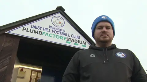 Aaron Hunt stands underneath a white sign for Daisy Hill Football club above the entrance to the clubhouse. He is wearing a woolly blue hat with clubs logo and a black hoodie.