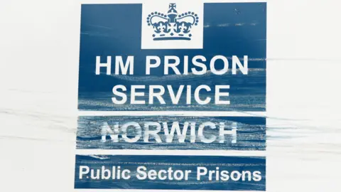 Martin Barber/BBC A blue and white sign at HMP Norwich, which has a crown logo on it. The sign reads: "HM Prison Service Norwich. Public sector prisons". It is slightly damaged.