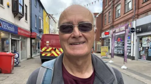 A balding man with sunglasses and a rucksack over a grey fleece, standing in Bangor town centre. It is a head and shoulders shot and he is looking at the camera. He has a grey fleece on and a maroon t-shirt and has a backpack on - as you can see the grey and blue straps.