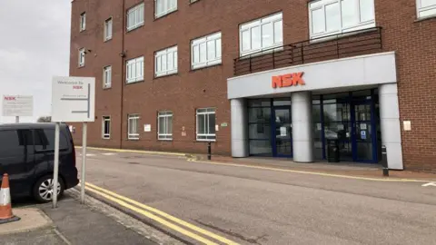 A brick building with large metal shutter in the middle. A sign at the top states NSK Newark plant.