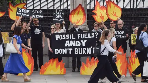 Getty Images Climate activists stand outside a wrought-iron fence holding flame-shaped placards and signs opposing North Sea oil and gas projects, including Rosebank and Jackdaw, while pedestrians walk past in the foreground.
