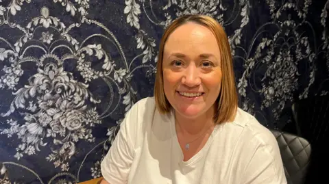 Lorna Myers sits against a backdrop of floral wallpaper. She is wearing a white t-shirt and has shoulder length light-brown hair. 