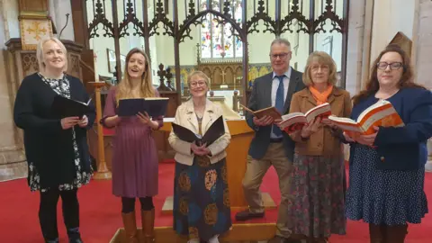 Sally Graham Five ladies, one man stand in a church with music books in their hands, behind them is a stained glass window and an alter