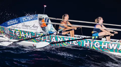 World's Toughest Row The picture shows two rowers powering through dark, choppy water in a brightly decorated ocean‑rowing boat. The boat has the words World’s Toughest Row on its cabin and colourful branding along the hull, including the name Team Atlantic Highway. 