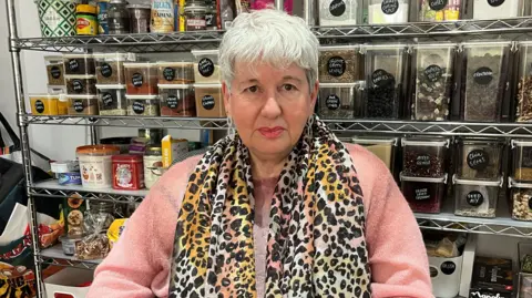 A woman wearing a pink top and leopard print scarf. She is in a shop with shelves behind her.