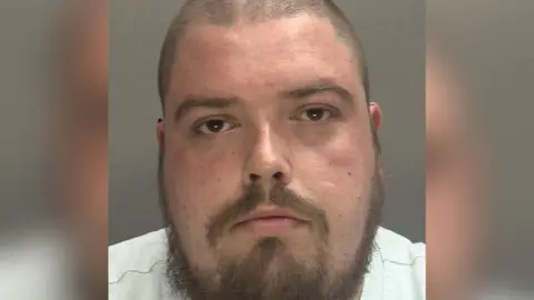 A police mugshot of Richard Britton, he has a shaved head and a beard.