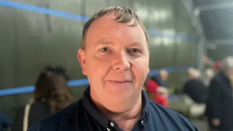 BBC/Laura Foster Andy Smith wears a navy blue polo shirt and looks into the camera. He is standing in a former air shelter which is being used as the venue for Dorothy Smith's birthday party