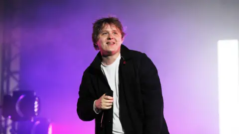 BBC Lewis Capaldi performs onstage - he is wearing a white shirt and black jacket, is holding a microphone and smiling
