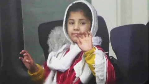 PA Media A child, wearing a white hood and red robe, waves from a bus after leaving the aircraft