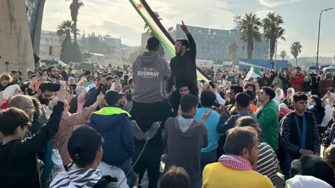 A celebratory crowd gathers outside the Opera House, clapping, arms raised, and taking videos, with two men on the shoulders of others, and some people waving Syrian opposition flags