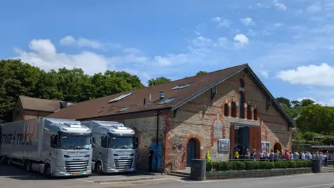 The Cheese and Grain frm the outside. It is a brick building with a big entrance. There are two stage trucks parked next to the building and a queue is leading inside the building.