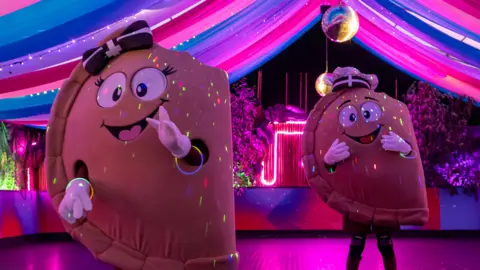 Two people wearing pasty costumes on a dance floor. The costumes have smiley faces on them. The dance floor is lit up with a purple light. Disco balls are hanging from the ceiling.
