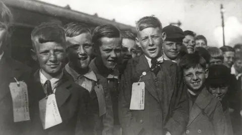 Lowestoft Central Project A black and white archive image of a group of young boys huddled together as they wait to board a train. They are all looking into the camera. They have paper identity tags attached to their clothing.