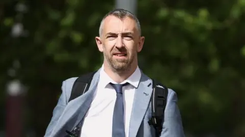 PA Media Daithí McKay is wearing a white shirt and a navy tie. He has a grey blazer on and a black rucksack. He has short grey hair and a beard.