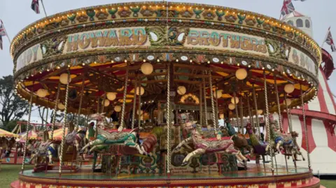 A brightly coloured vintage carousel decorated with lights and painted horses is set up on grass at a fairground, with “Hayward Brothers” written across the top. Union Jack flags fly above the ride, and a large striped tent stands in the background, adding to the traditional fair atmosphere.
