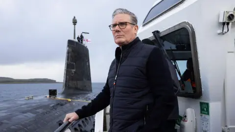 Keir Starmer on a boat next to a grey nuclear submarine. He is wearing a blue gilet over a blue or black jumper. He has grey hair and is wearing glasses. There are two men standing on the conning tower of the submarine