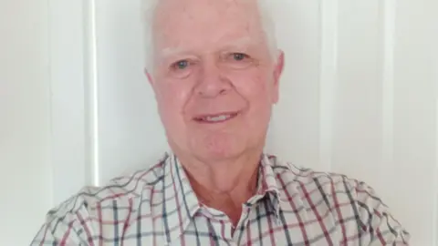 Rob Aldenton Rob Aldenton pictured. He is wearing a checkered shirt which has blue and red lines on. He has grey hair and is looking towards the camera and smilng. 