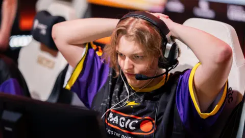 Esports Foundation A competitor at the Esports World Cup watches a screen intently with their hands clasped behind their head, creating an impression of anticipation or nervousness. They wear a large gaming headset with a microphone and a black and purple team jersey.