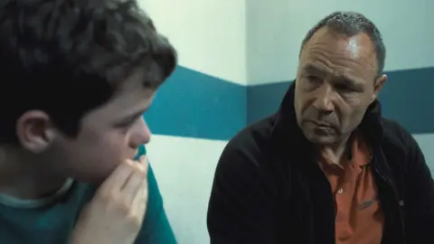 A still from a scene in Adolescence featuring a young boy talking to his father inside a prison cell. The boy has dark hair and is looking towards his dad, who wears a concerned expression. He has short grey hair.