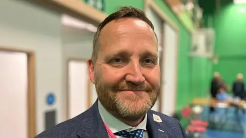 Councillor Matt Dormer is pictured - he is inside a leisure centre building wearing a blue suit jacket and a green shirt, with a lanyard around his neck.