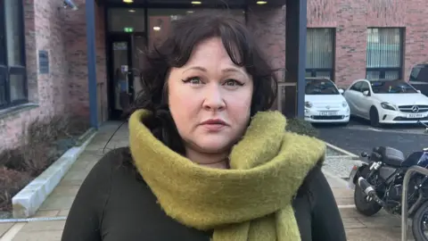 Sara Morrison with short dark hair looks at the camera. She is wearing a pale green scarf and is standing outside in front of a building and a number of parked cars. 