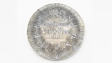 John Rolfe Auctions Reverse of Medal. A series of spiky shaped flowers adorn the smooth edges of the medal, with the words Eurovision Grand Prix de la Chanson 1967, Puppet on a String written on it.