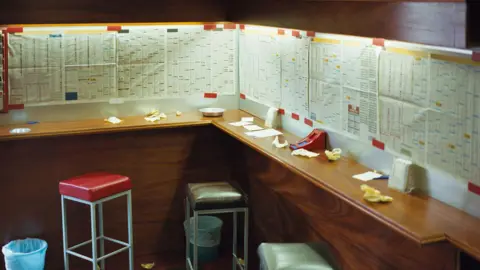 Getty Images Stock image of the inside of a betting shop, the walls are covered with race listings and there is a bench with stools. Numerous paper slips are discarded around the room. 