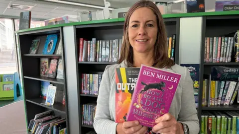A woman stands in a library in front of a large book case containign rows of dozens of book. She is holding two books in her hands, one has a magenta cover and the other is orange, black and yellow.