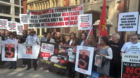PA Bereaved relatives affected by mental health deaths in Essex stand outside Arundel House in London. They are holding posters and photographs of loved ones that have died. One is also holding an Essex flag. One banner reads: "If mental health trusts in Essex had learned from mistakes, there would be one death instead of thousands of deaths"