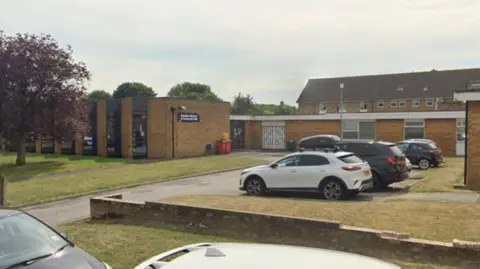 A Google Streetview image of a brown brick library building on a street with grass and trees in front of it. A few cars are parked and flats are in the background.