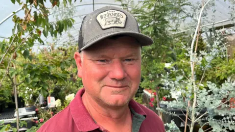 A man has a grey baseball cap on his head and a mauve open collar t-shirt top on, behind him are plants inside a polytunnel.