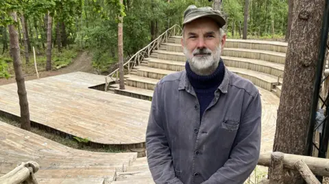Darren Rozier/BBC A man stands above an outdoor theatre. He wears a green cap, a faded blue shirt with a navy blue turtle neck jumper underneath. He has a grey beard and rests his hands in front of his body as he smiles at the camera.