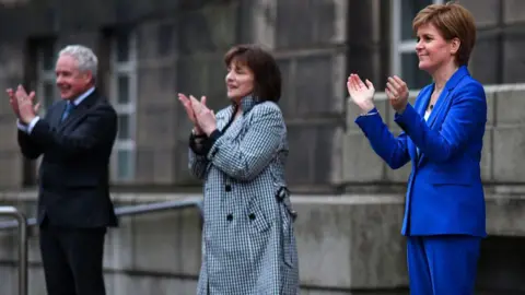 Getty Images Malcolm Wright, a man in a dark suit; Jeane Freeman, a woman in a black and white checked coat and Nicola Sturgeon, a woman in an electric blue trouser suit, stand in a row applauding outside a grey stone building (St Andrew's House).