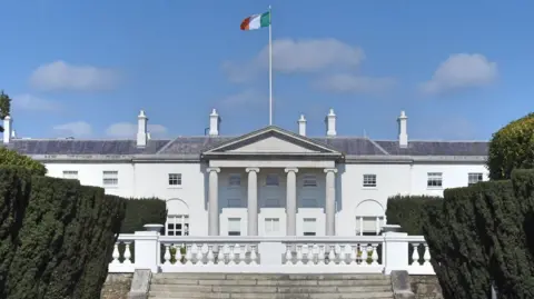Getty Images The exterior of Áras an Uachtaráin - the official residence of the Irish president. It is a white building, not dissimilar to the White House in Washington. It features four large columns at the front of it. An Irish tricolour is flown from a flagpole on the roof of the building.